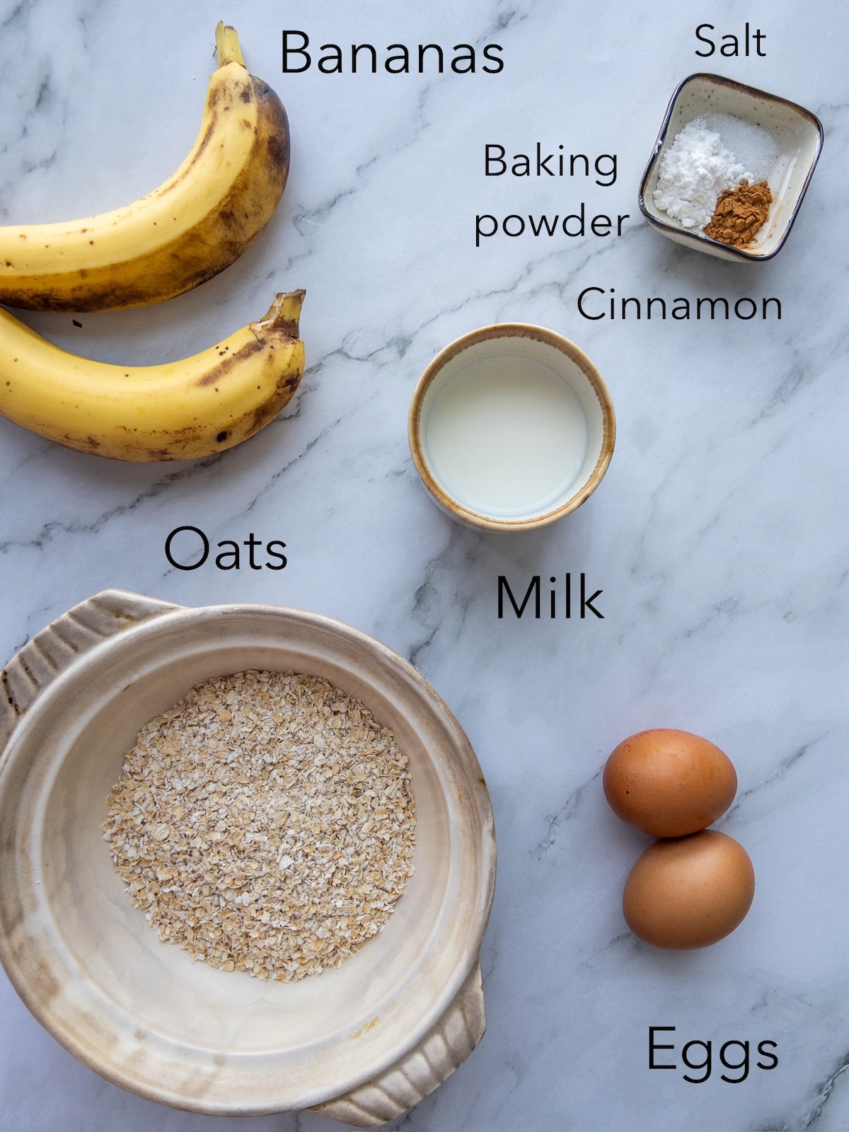 Ingredients for banana pancakes presented on a table. Includes oats, eggs, milk and bananas
